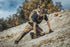 Wearing Condors Paladin Tactical Pants, a person with a backpack kneels on rocky, sloped terrain with trees and a cloudy sky in the background.